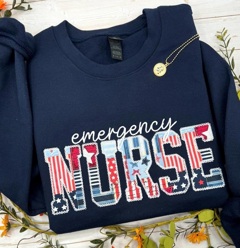 Op de afbeelding: Een marineblauw sweatshirt met ronde hals en de witte tekst "emergency" boven het woord "NURSE" in grote blokletters. De letters zijn gevuld met een patriottisch rood, wit en blauw patroon van sterren en strepen.