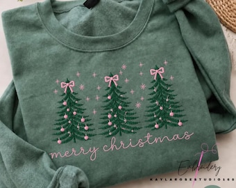 Op de afbeelding: Een heather groen sweatshirt met ronde hals voorzien van een geborduurd ontwerp van drie donkergroene kerstbomen versierd met roze ornamenten en roze strikken, inclusief de tekst merry christmas in roze schrift.