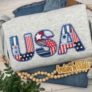 May include: A light gray sweatshirt featuring the letters USA appliqued in a patchwork style. The letters are constructed from fabric patterns including red and white stripes, blue with white stars, and red with white stars, all outlined with blue stitching.