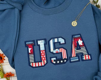 Può includere: Una felpa blu con la scritta "USA" applicata sul davanti con un design patchwork patriottico. Le lettere presentano motivi rossi, bianchi e blu con stelle e strisce. Una collana d'oro con un ciondolo rotondo poggia sulla felpa.