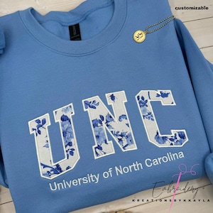 May include: A light blue sweatshirt with the letters "UNC" in a floral pattern, with the words "University of North Carolina" below. A gold-colored charm hangs from the neckline. The word "customizable" is in the top right corner.