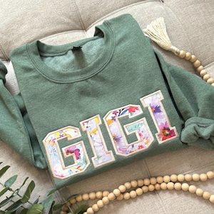 以下が含まれることがあります： 「GIGI」の文字が大きく花柄でデザインされたグリーンのスウェットシャツ。文字はクリーム色の縁取り。柔らかい素材でできており、カジュアルウェアに適しています。