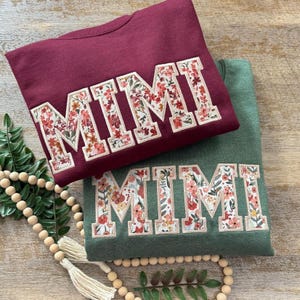 May include: Two sweatshirts with the word "MIMI" spelled out in large, white letters with a floral pattern. The sweatshirts are maroon and forest green.