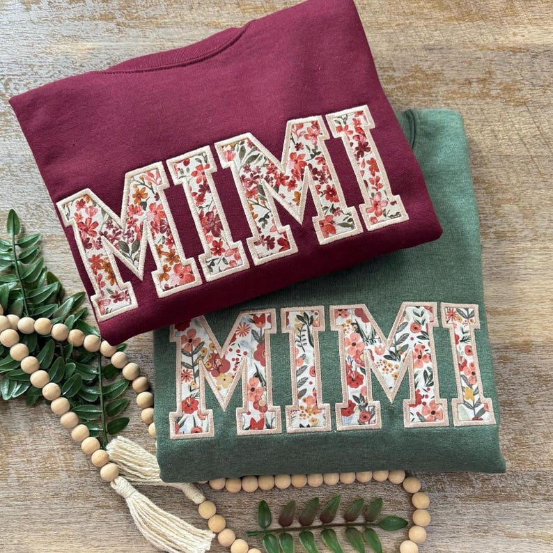 May include: Two sweatshirts with the word "MIMI" spelled out in large, white letters with a floral pattern. The sweatshirts are maroon and forest green.