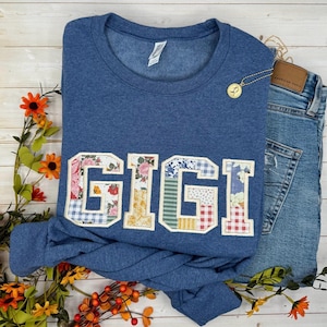 以下が含まれることがあります： パッチワークのデザインで「GIGI」の文字がアップリケされた青いスウェットシャツ。文字は花柄、ギンガムチェック、その他の模様の生地を使用。金のネックレスとジーンズがスウェットシャツの隣にあります。