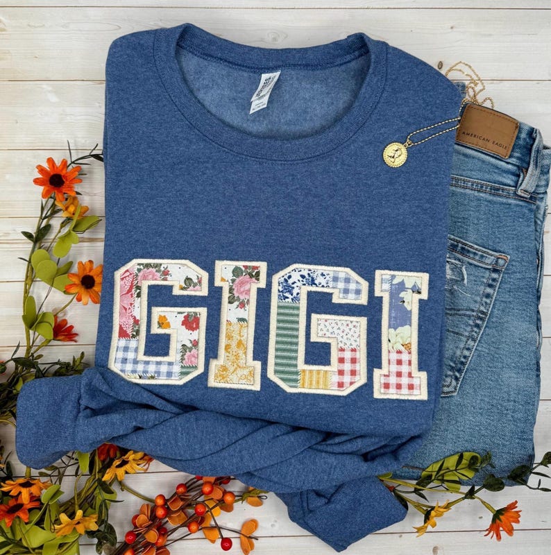以下が含まれることがあります： パッチワークのデザインで「GIGI」の文字がアップリケされた青いスウェットシャツ。文字は花柄、ギンガムチェック、その他の模様の生地を使用。金のネックレスとジーンズがスウェットシャツの隣にあります。