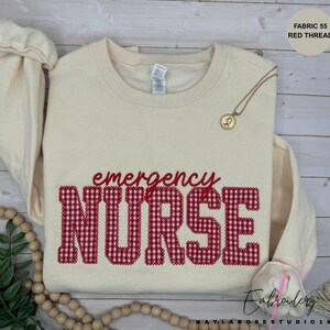 May include: A cream-colored crewneck sweatshirt featuring the word NURSE in large, red and white gingham-patterned block letters, with the word emergency written in smaller red script above it. A gold pendant necklace rests on the collar.