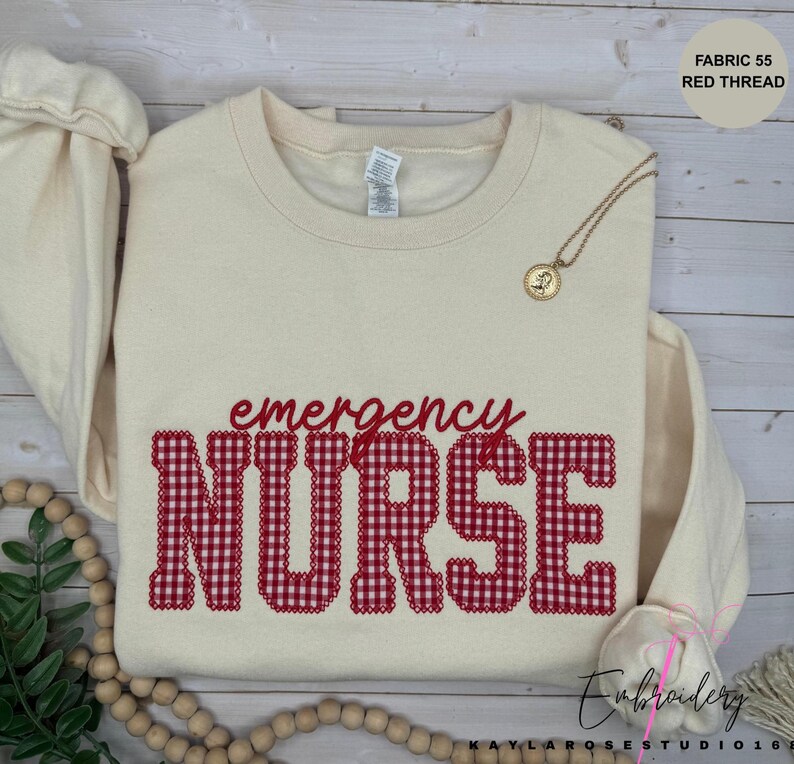May include: A cream-colored crewneck sweatshirt featuring the word NURSE in large, red and white gingham-patterned block letters, with the word emergency written in smaller red script above it. A gold pendant necklace rests on the collar.