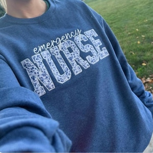 May include: A blue sweatshirt with the words "emergency NURSE" in white, the word "NURSE" is filled with a floral pattern. The sweatshirt is a solid blue colour. The image is taken outdoors.