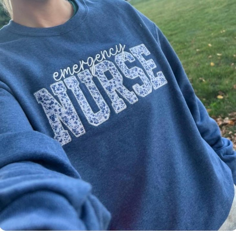 May include: A blue sweatshirt with the words "emergency NURSE" in white, the word "NURSE" is filled with a floral pattern. The sweatshirt is a solid blue colour. The image is taken outdoors.
