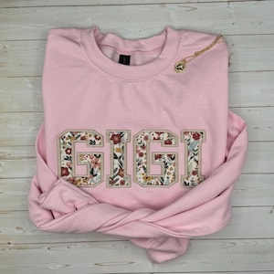 以下が含まれることがあります： フローラルパターンで「GIGI」と刺繍された、明るいピンク色のスウェットシャツ。ペンダント付きのゴールドネックレスがスウェットシャツの上にあります。衣類は明るい木製の表面に折りたたまれています。