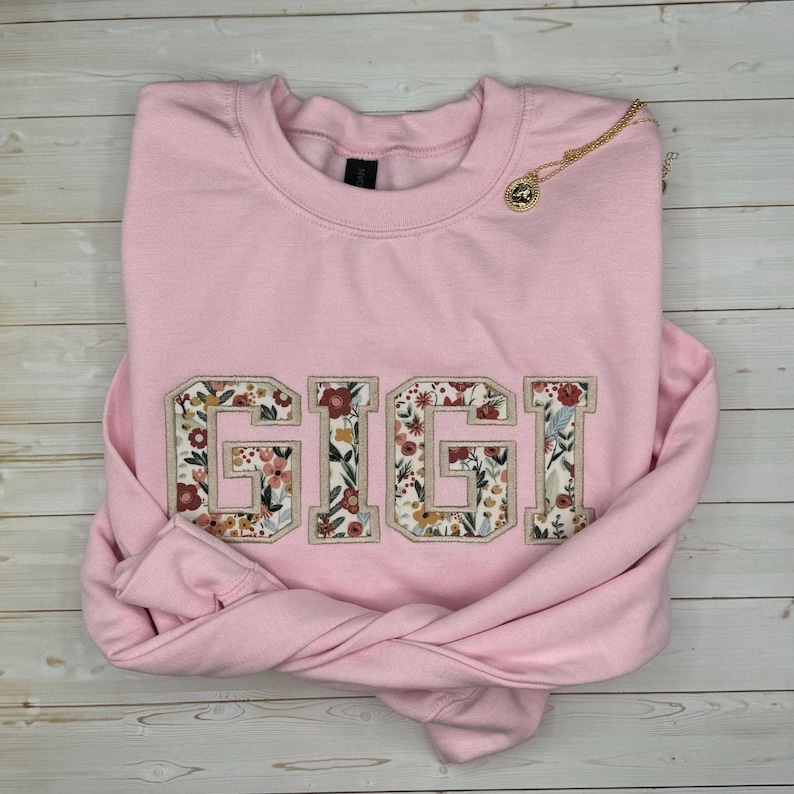 以下が含まれることがあります： フローラルパターンで「GIGI」と刺繍された、明るいピンク色のスウェットシャツ。ペンダント付きのゴールドネックレスがスウェットシャツの上にあります。衣類は明るい木製の表面に折りたたまれています。