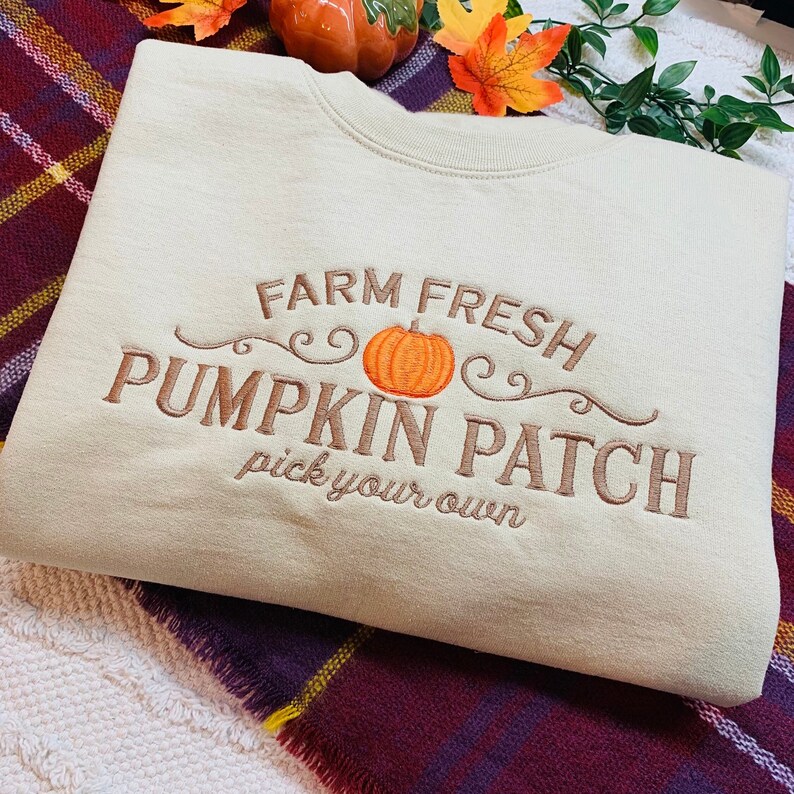 May include: A beige sweatshirt with an embroidered design that says "Farm Fresh Pumpkin Patch pick your own" with a small orange pumpkin in the centre.