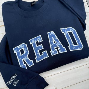 May include: A navy blue sweatshirt featuring the word "READ" in large, blue floral print letters. The sleeve has the embroidered text "Miss Rilee" with a small book design. The sweatshirt is folded on a light wood surface.