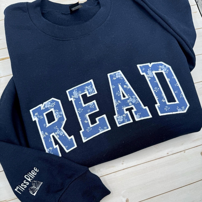 May include: A navy blue sweatshirt featuring the word "READ" in large, blue floral print letters. The sleeve has the embroidered text "Miss Rilee" with a small book design. The sweatshirt is folded on a light wood surface.