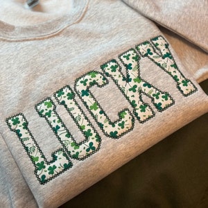 May include: A gray sweatshirt with the word "LUCKY" embroidered in green shamrock-patterned fabric.