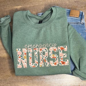 May include: Sage green sweatshirt with the words "emergency NURSE" in a floral design. The sweatshirt is paired with blue jeans. The floral design includes pink, orange, and yellow flowers.