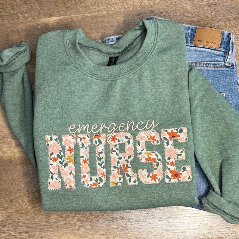 May include: Sage green sweatshirt with the words "emergency NURSE" in a floral design. The sweatshirt is paired with blue jeans. The floral design includes pink, orange, and yellow flowers.