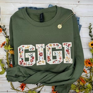 以下が含まれることがあります： 「GIGI」の文字が大きく花柄でデザインされたオリーブグリーンのスウェットシャツ。ペンダント付きのゴールドカラーのネックレスがスウェットシャツの上にかけられています。スウェットシャツはジーンズの上に置かれています。