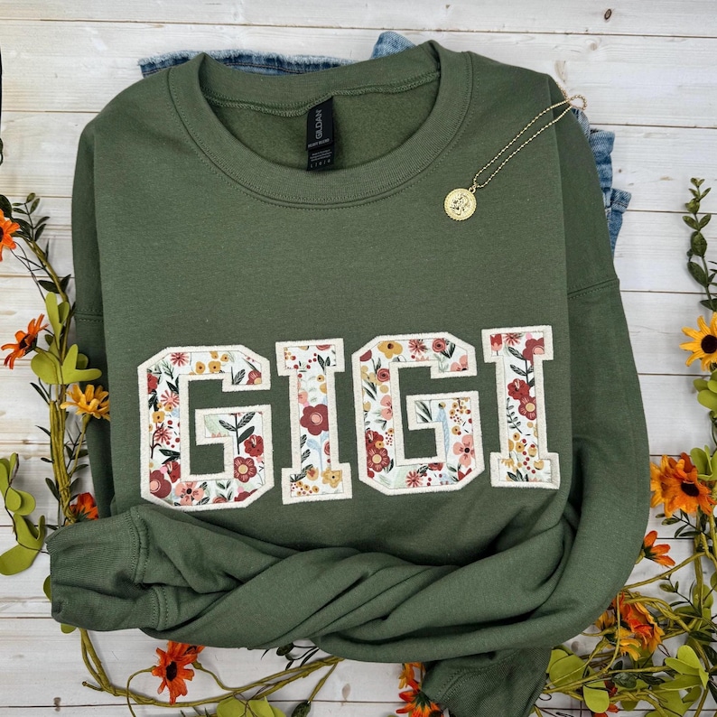 以下が含まれることがあります： 「GIGI」の文字が大きく花柄でデザインされたオリーブグリーンのスウェットシャツ。ペンダント付きのゴールドカラーのネックレスがスウェットシャツの上にかけられています。スウェットシャツはジーンズの上に置かれています。