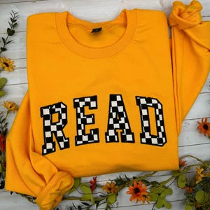 May include: A mustard yellow sweatshirt featuring the word "READ" in bold, chequered black and white letters. The garment is folded, showing the collar and sleeves. Floral decorations are arranged around the sweatshirt.