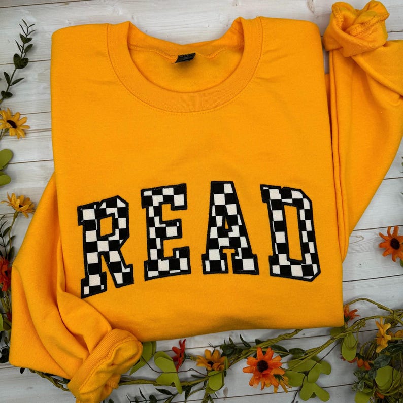 May include: A mustard yellow sweatshirt featuring the word "READ" in bold, chequered black and white letters. The garment is folded, showing the collar and sleeves. Floral decorations are arranged around the sweatshirt.