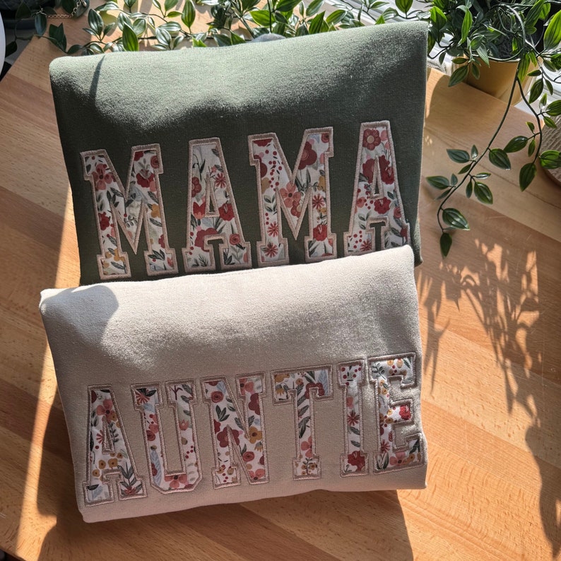 May include: Two sweatshirts, one olive green and one beige, are displayed. The word "MAMA" is embroidered on the green sweatshirt, and "AUNTIE" is embroidered on the beige sweatshirt. The letters are filled with a floral pattern.