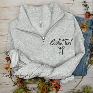 May include: A light gray quarter-zip sweatshirt with the words "Custom Text" and a bow graphic in black. The sweatshirt is folded on a wooden surface with denim jeans and floral accents.