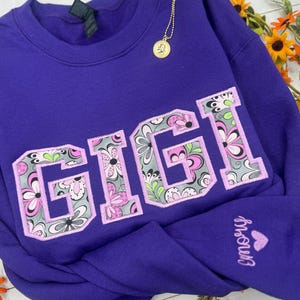 以下が含まれることがあります： 大きな花柄の文字で「GIGI」と書かれた紫色のスウェットシャツ。スウェットシャツはクルーネックと長袖です。前面にはペンダント付きのゴールドネックレスがぶら下がっています。袖には「Emory」という名前がハートで刺繍されています。