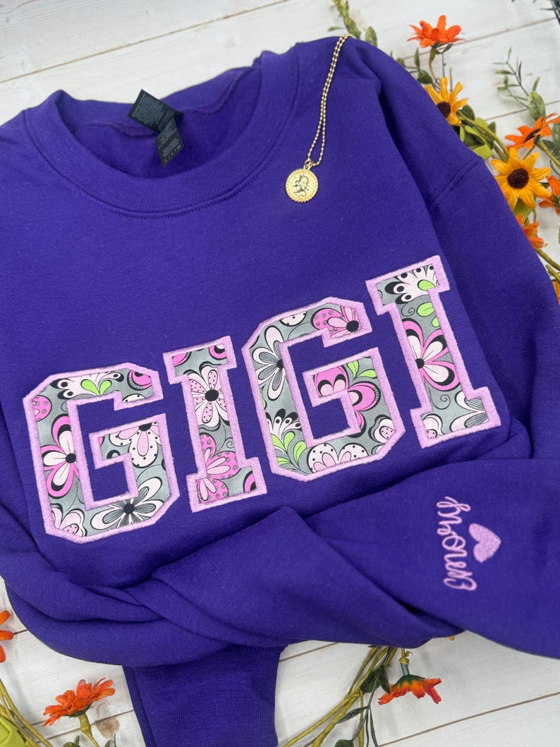 以下が含まれることがあります： 大きな花柄の文字で「GIGI」と書かれた紫色のスウェットシャツ。スウェットシャツはクルーネックと長袖です。前面にはペンダント付きのゴールドネックレスがぶら下がっています。袖には「Emory」という名前がハートで刺繍されています。