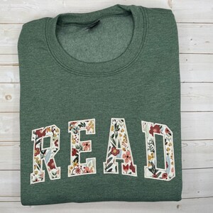 May include: A green sweatshirt featuring the word "READ" in large, floral-patterned letters. The letters are white with a colourful design of flowers and leaves. The sweatshirt is folded neatly on a light wood surface.