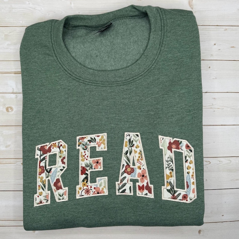May include: A green sweatshirt featuring the word "READ" in large, floral-patterned letters. The letters are white with a colourful design of flowers and leaves. The sweatshirt is folded neatly on a light wood surface.