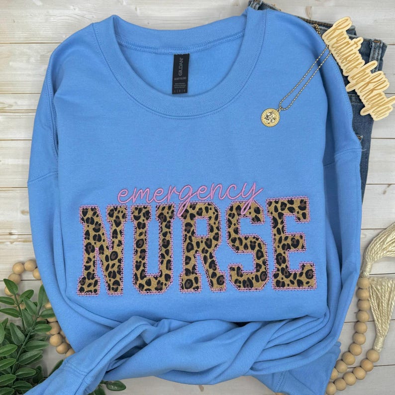 Op de afbeelding: Een lichtblauw sweatshirt met ronde hals en het woord NURSE in grote blokletters met luipaardprint en een roze omlijning. Boven het woord staat emergency in roze cursief schrift. Een gouden hangerketting ligt op de stof.
