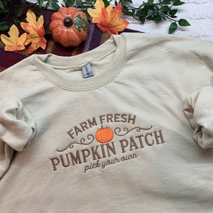 May include: A beige sweatshirt with an embroidered design that says "Farm Fresh Pumpkin Patch pick your own" with a small orange pumpkin in the centre.
