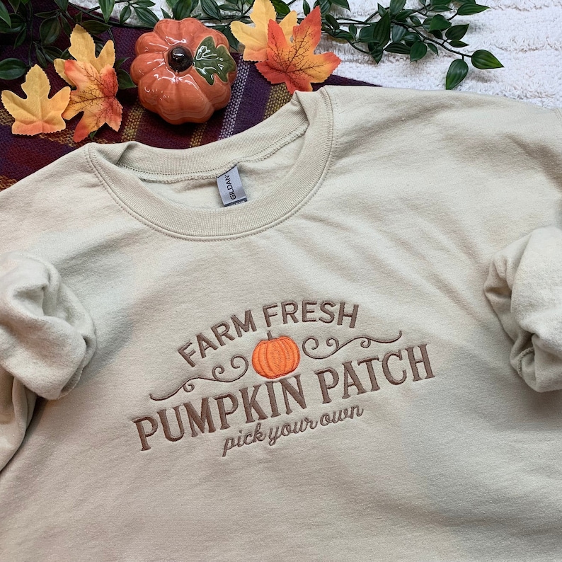 May include: A beige sweatshirt with an embroidered design that says "Farm Fresh Pumpkin Patch pick your own" with a small orange pumpkin in the centre.