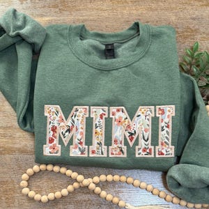 May include: A green sweatshirt with the word "MIMI" embroidered in white letters with a floral pattern. The letters are outlined in pink.