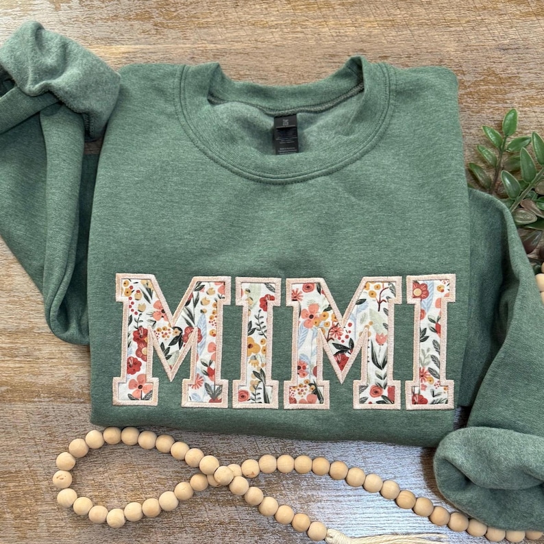 May include: A green sweatshirt with the word "MIMI" embroidered in white letters with a floral pattern. The letters are outlined in pink.