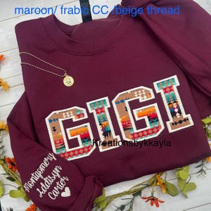 以下が含まれることがあります： 鮮やかな南西部のパターンで「GIGI」という文字がプリントされたマルーン色のスウェットシャツ。袖には「Montgomery Addison Carter」という名前が白で刺繍されています。ペンダント付きのゴールドネックレスがスウェットシャツの上にかけられています。