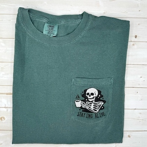 May include: A teal Comfort Colors t-shirt with a pocket featuring an embroidered skeleton holding a coffee cup. The text "STAYING ALIVE" is below the skeleton. The shirt is folded on a light wood surface.