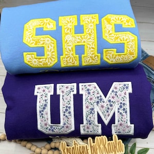 May include: Two sweatshirts, one light blue and one purple, are stacked. The light blue sweatshirt has the letters "SHS" in yellow with a floral pattern. The purple sweatshirt has "UM" in white with a floral pattern.
