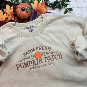 May include: A beige sweatshirt with the text "Farm Fresh Pumpkin Patch pick your own" embroidered in brown on the front. The text is surrounded by a decorative swirl design and a small orange pumpkin.