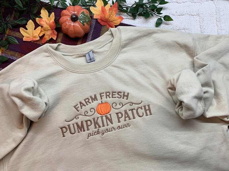May include: A beige sweatshirt with the text "Farm Fresh Pumpkin Patch pick your own" embroidered in brown on the front. The text is surrounded by a decorative swirl design and a small orange pumpkin.