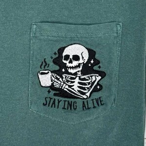 May include: Green t-shirt with a pocket featuring a black and white embroidered skeleton holding a coffee cup. The text "STAYING ALIVE" is below the skeleton.