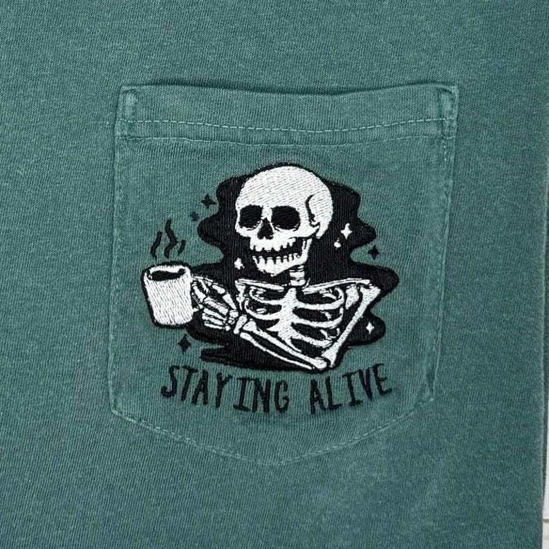 May include: Green t-shirt with a pocket featuring a black and white embroidered skeleton holding a coffee cup. The text "STAYING ALIVE" is below the skeleton.