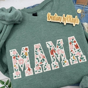 May include: Heather green sweatshirt with the word "MAMA" in large, floral-patterned letters. The sweatshirt has a crew neck and long sleeves. The text "Kreations by R Kayla" is on a wooden tag.