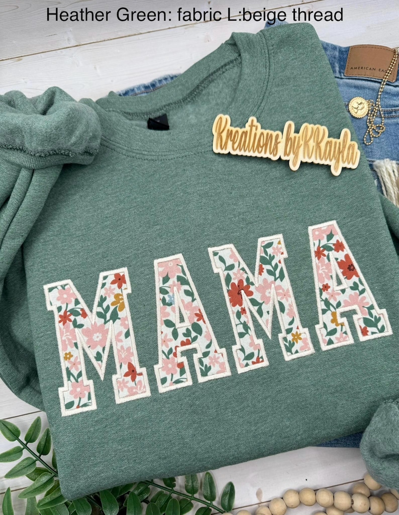 May include: Heather green sweatshirt with the word "MAMA" in large, floral-patterned letters. The sweatshirt has a crew neck and long sleeves. The text "Kreations by R Kayla" is on a wooden tag.