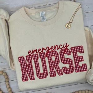 May include: A cream-colored crewneck sweatshirt featuring the word emergency in red cursive script above the word NURSE in large, block-style letters with a red and white gingham pattern. A gold-toned circular pendant necklace rests on the fabric.