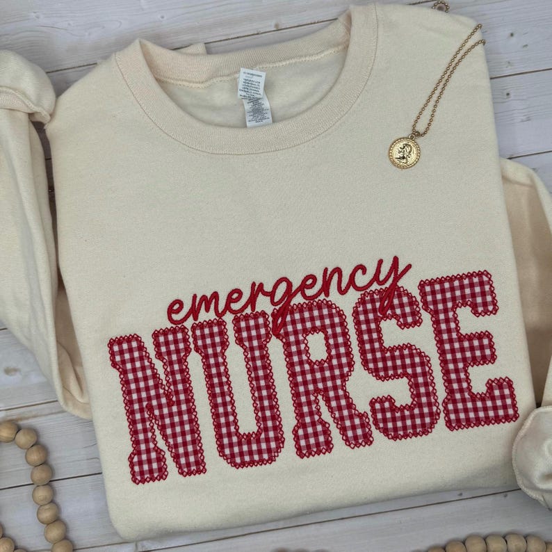 May include: A cream-colored crewneck sweatshirt featuring the word emergency in red cursive script above the word NURSE in large, block-style letters with a red and white gingham pattern. A gold-toned circular pendant necklace rests on the fabric.
