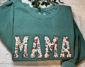 May include: A sage green crewneck sweatshirt featuring the word MAMA appliqued across the chest. The letters are filled with a cream-colored fabric displaying a floral pattern of red, orange, and green, outlined with dark green stitching.