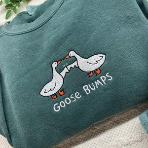 K&ouml;nnte beinhalten: Ein tealgr&uuml;ner Pullover mit einem gestickten Design aus zwei G&auml;nsen, die sich gegen&uuml;berstehen, mit dem Text "Goose Bumps" darunter.